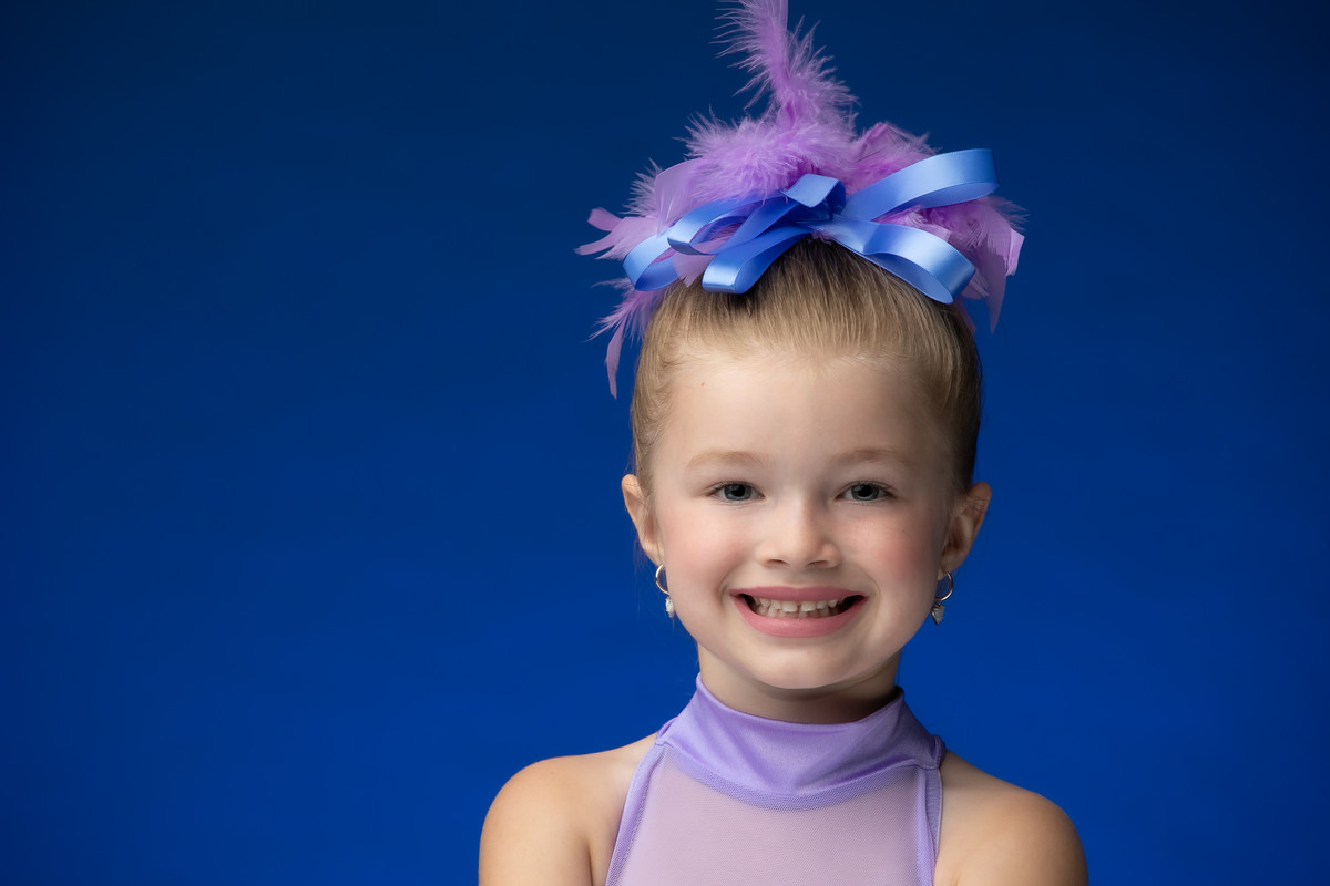 Close-up portrait of a young dancer in lavender with a feather hairpiece on a blue backdrop